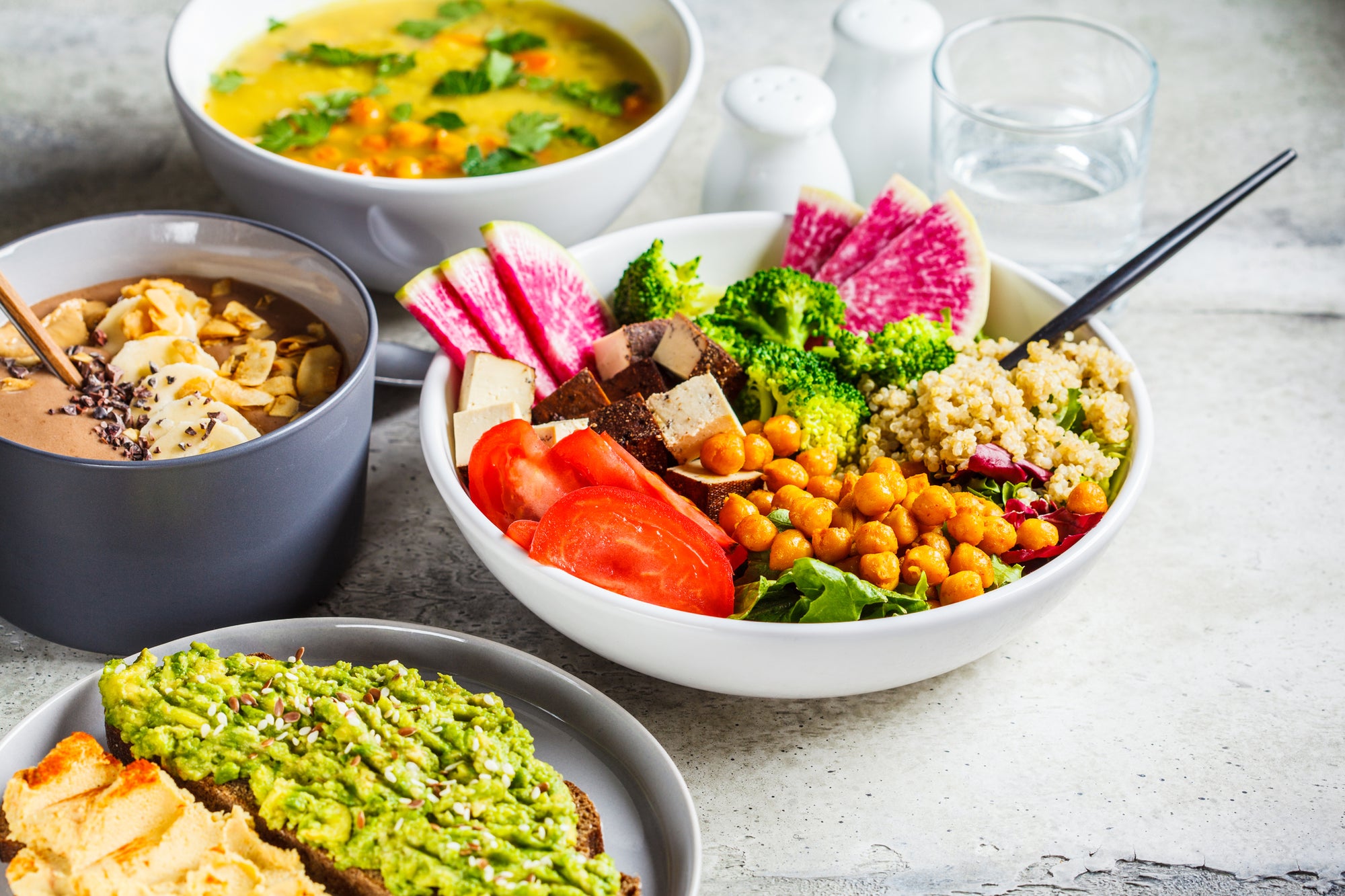 Bowls of plant-based food, fruits & vegetables such as chickpeas, tofu, guacamole, soup, and broccoli